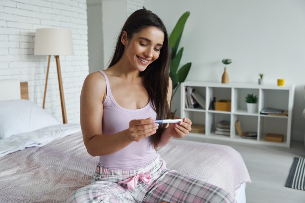 Happy young woman looking at pregnancy test while sitting on bed