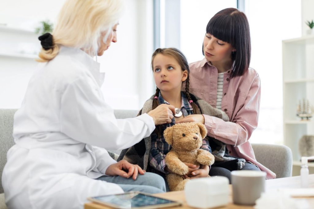 A female doctor uses a stethoscope to listen to a young girl's heart while her mother comforts her. The girl holds a teddy bear.