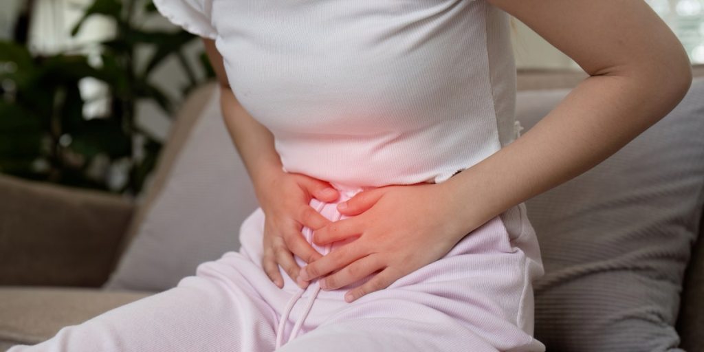Woman Experiencing Stomach Pain While Sitting on a Couch in a Home Setting, Clutching Her Abdomen in Discomfort