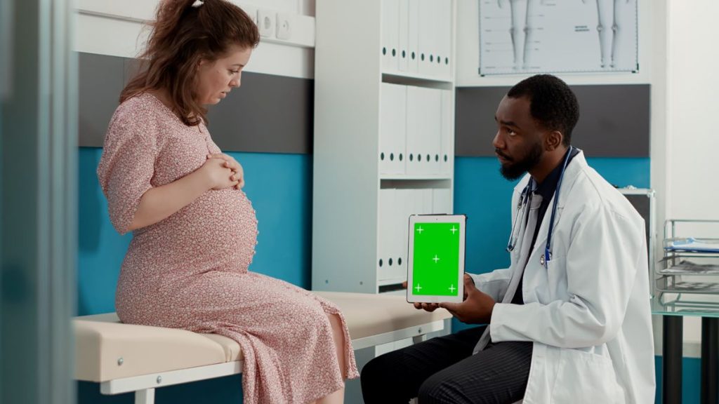 Male physician showing digital tablet with greenscreen display to pregnant patient on bed