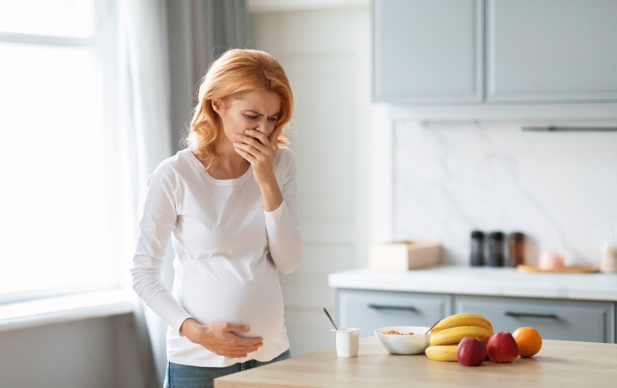 A pregnant woman stands in a modern kitchen, feeling nauseous while holding her stomach